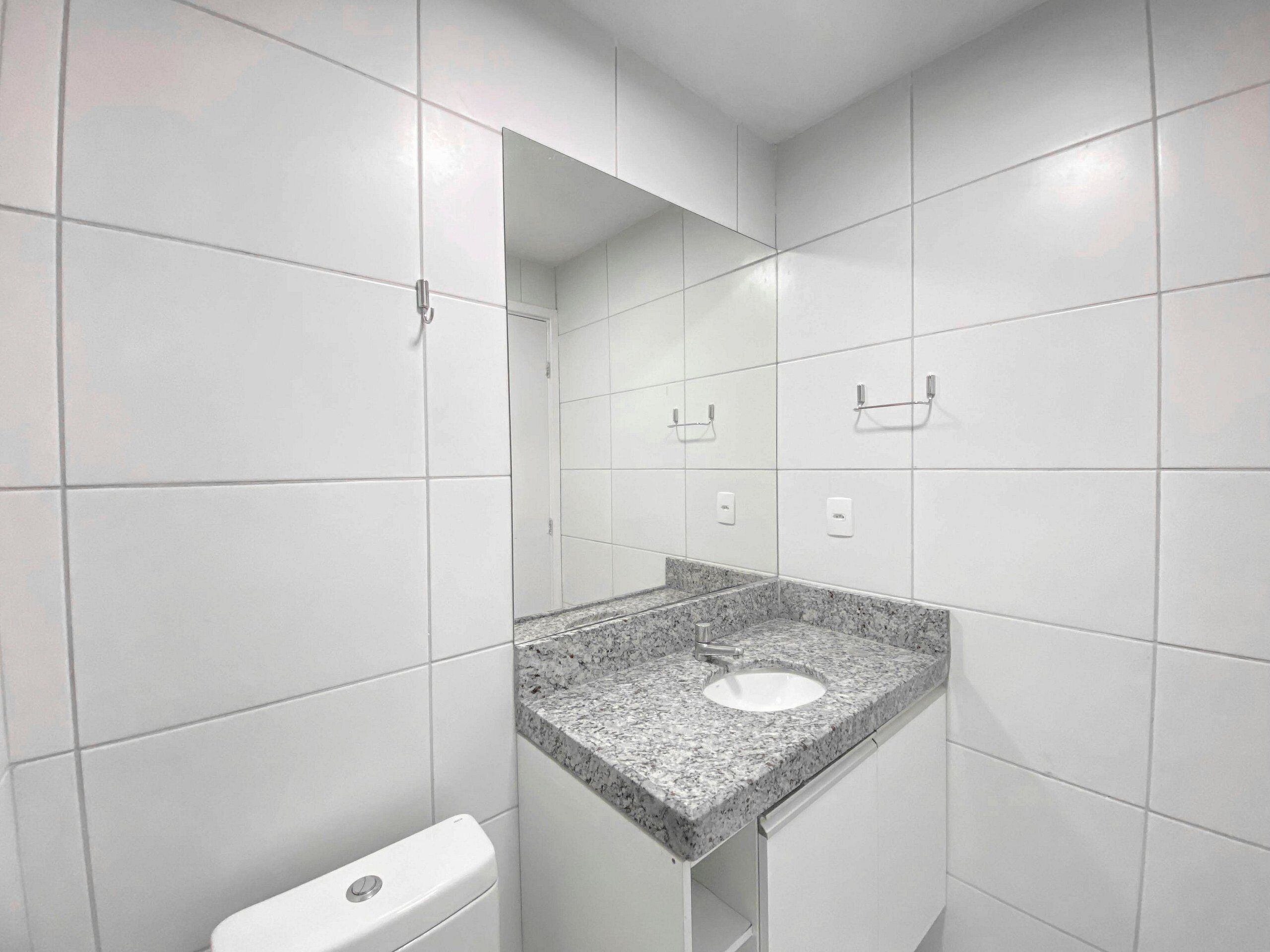 Ein Badezimmer mit weißen Fliesen, einem Waschbecken mit Granitplatte, einer Toilette und einem Spiegel.