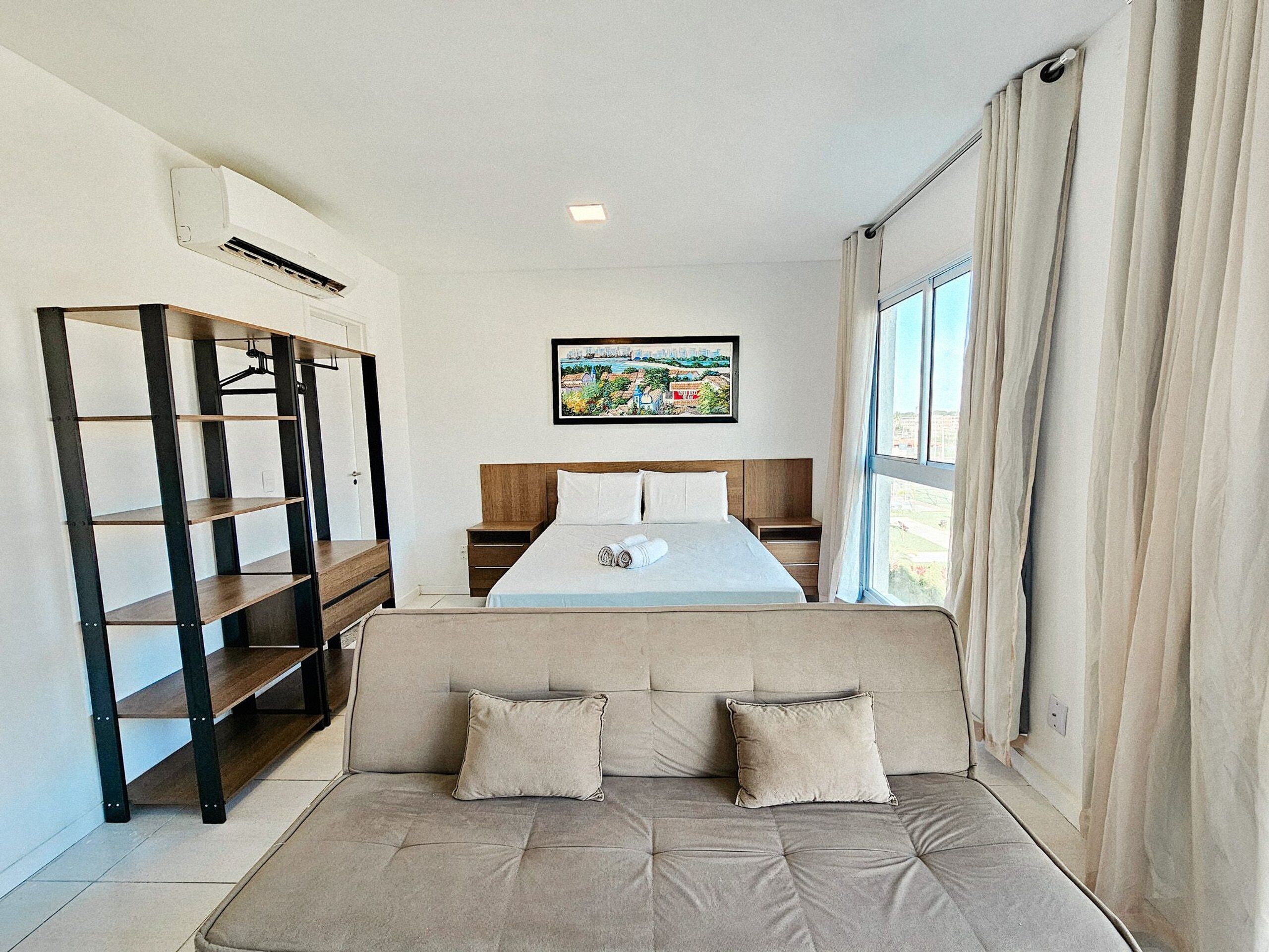A hotel room with a bed, a sofa, a bookshelf, a painting, and a window with curtains.