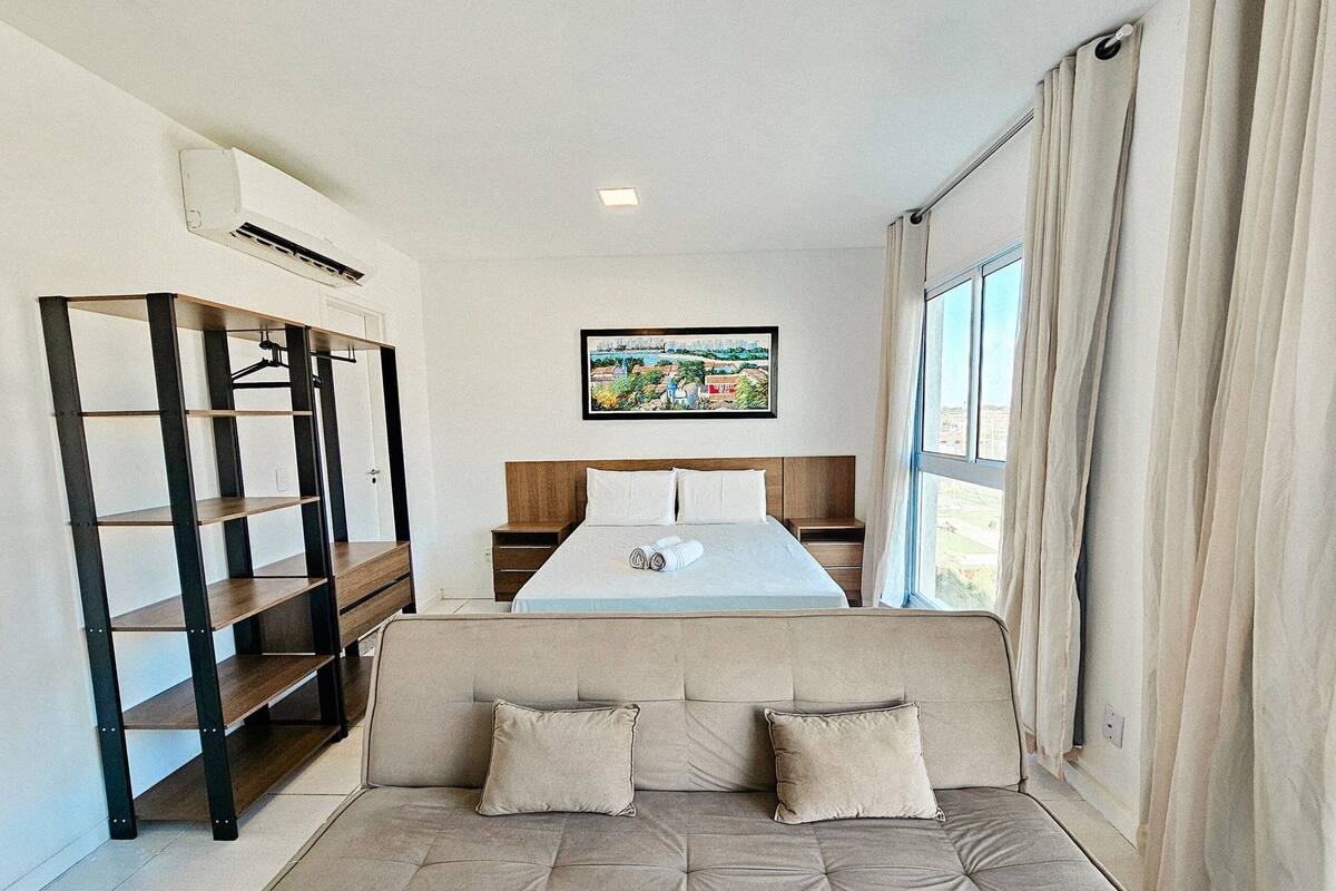 A hotel room with a bed, a sofa, a bookshelf, a painting, and a window with curtains.
