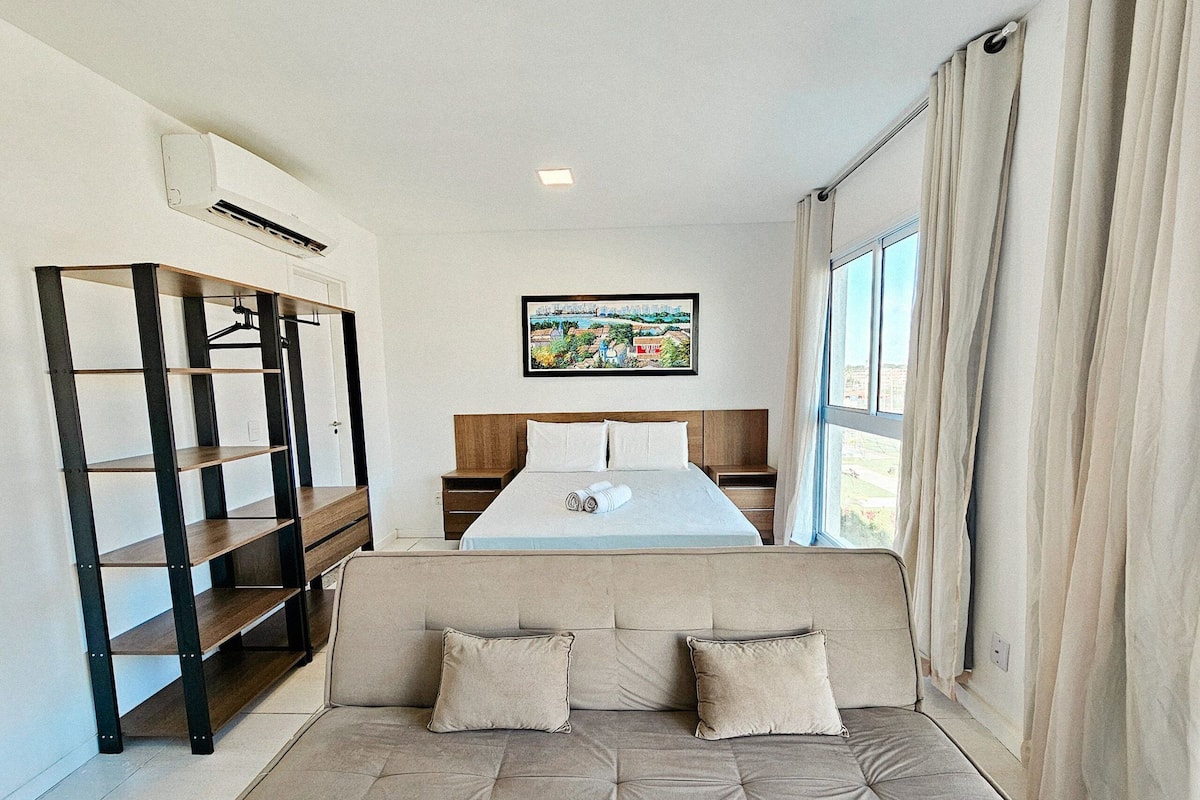 A hotel room with a bed, a sofa, a bookshelf, a painting, and a window with curtains.