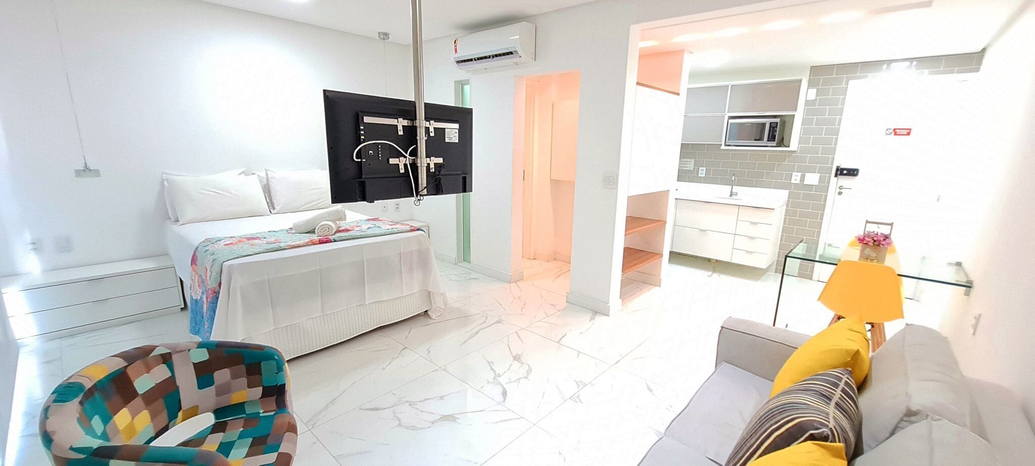 Una habitación de hotel moderna con una cama, un sofá, un sillón colorido, una zona de cocina con microondas y un ventanal de vidrio que da a un balcón.