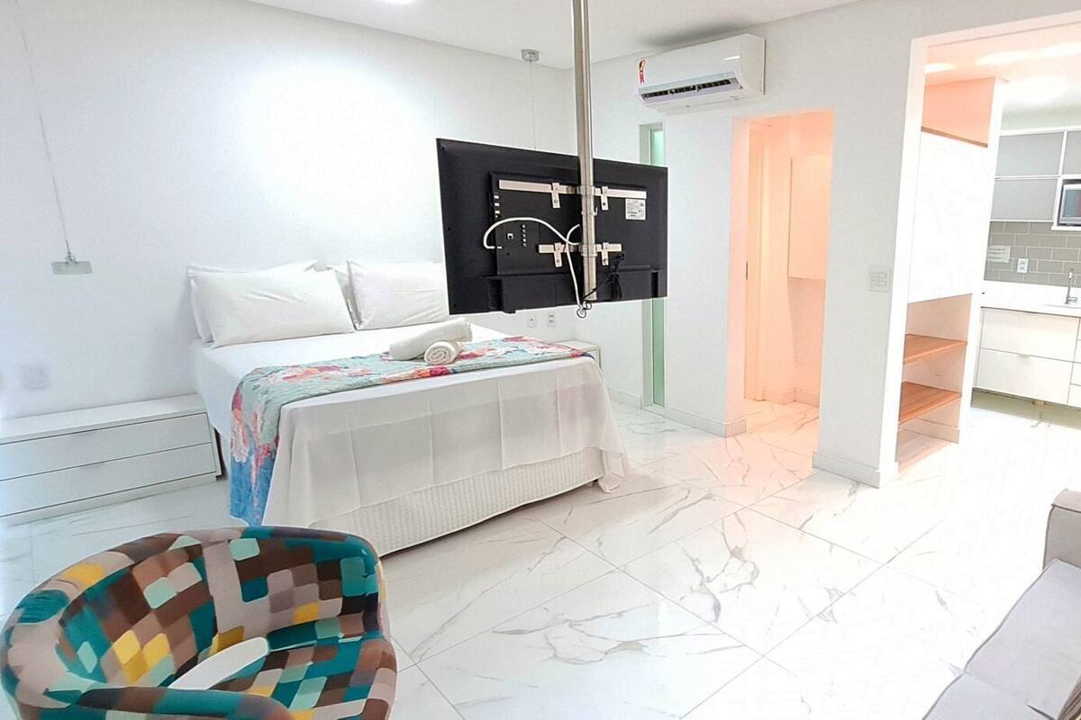 A modern hotel room with a bed, a sofa, a colorful armchair, a kitchen area with a microwave, and a glass door leading to a balcony.