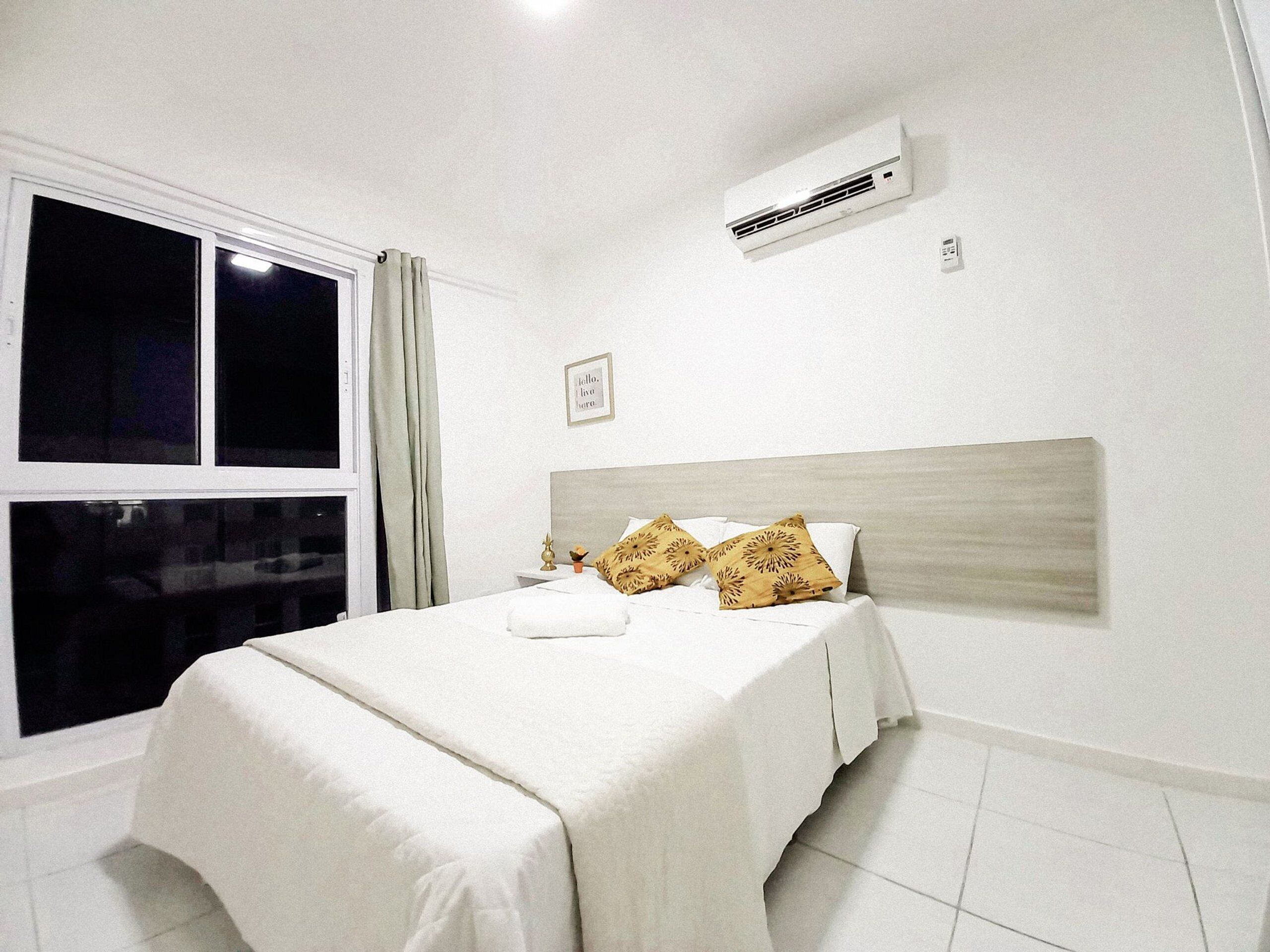 Un dormitorio con cama, ropa de cama blanca, una ventana con cortinas, un equipo de aire acondicionado y un cuadro pequeño en la pared.