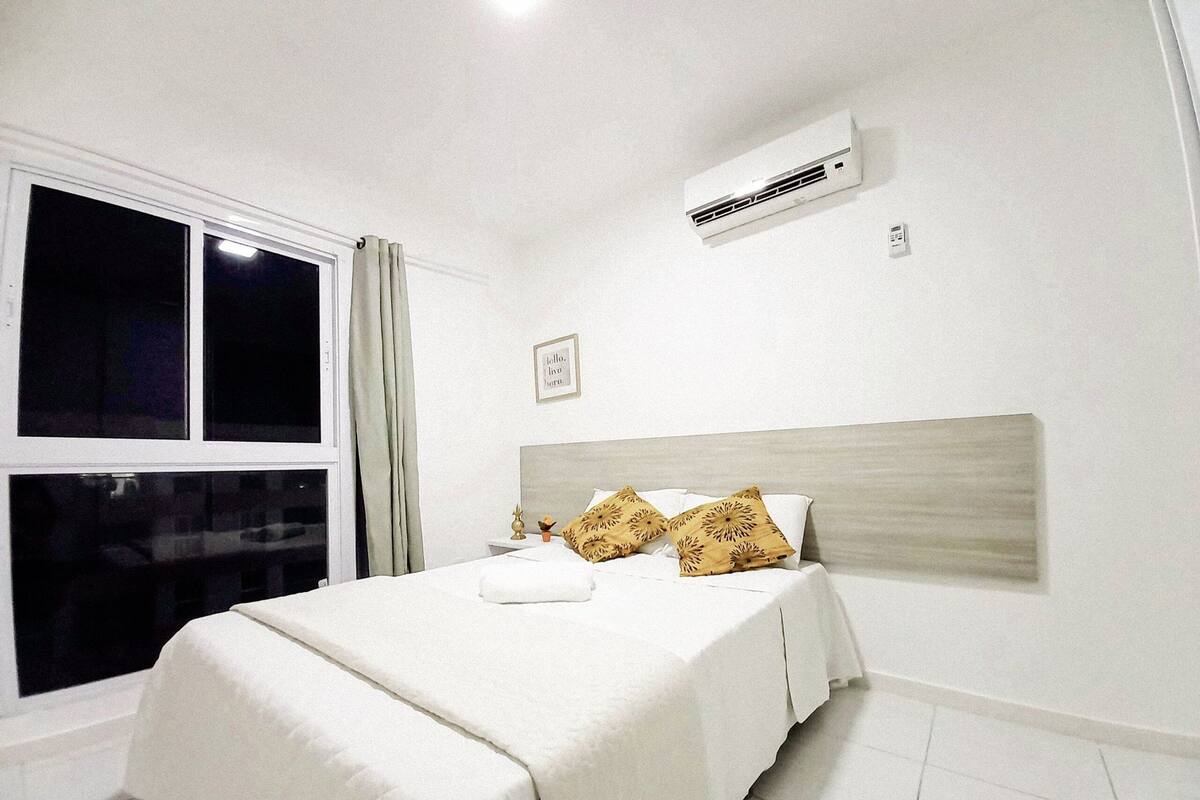 A bedroom with a bed, white bedding, a window with curtains, an air conditioning unit, and a small picture on the wall.