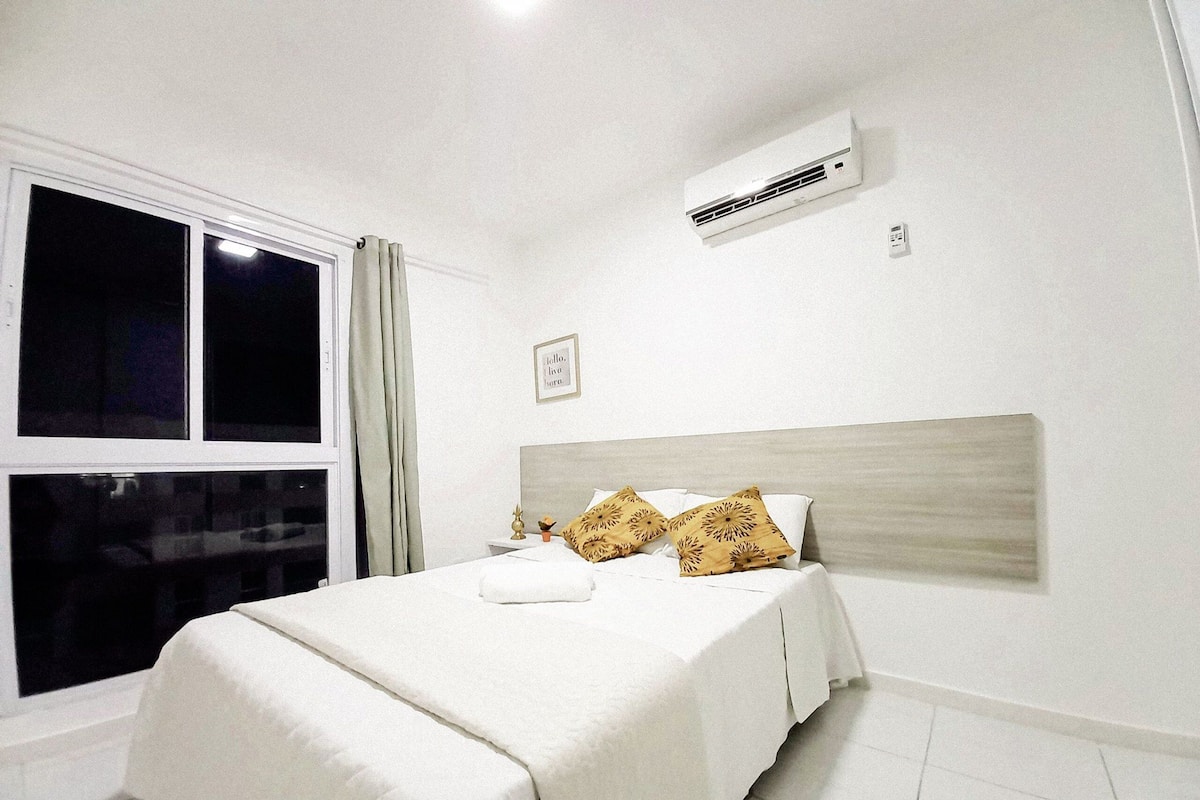 Un dormitorio con cama, ropa de cama blanca, una ventana con cortinas, un equipo de aire acondicionado y un cuadro pequeño en la pared.