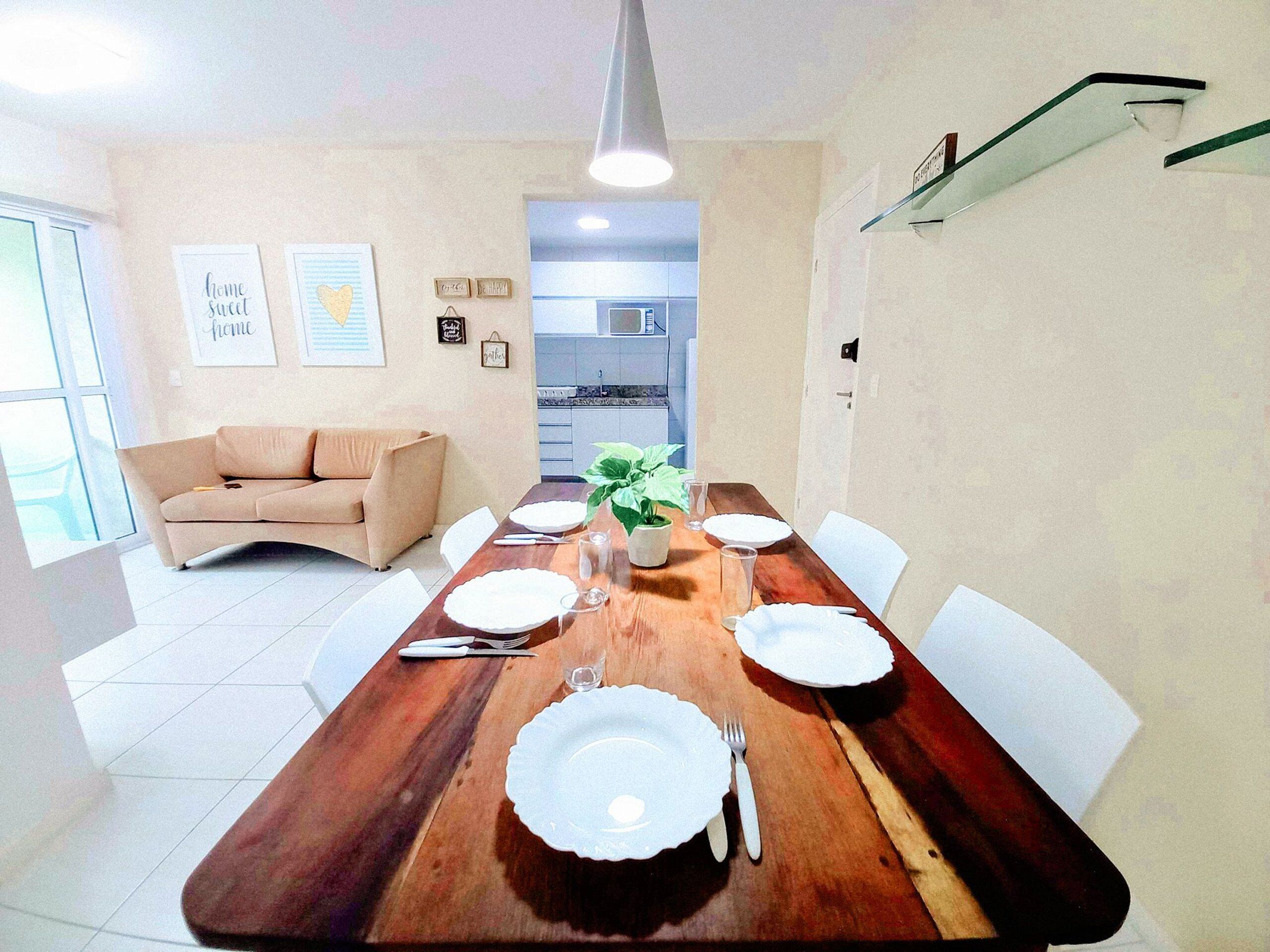 A dining area with a wooden table set for four, surrounded by white chairs. A beige sofa is positioned against the wall, and a kitchen is visible in the background.