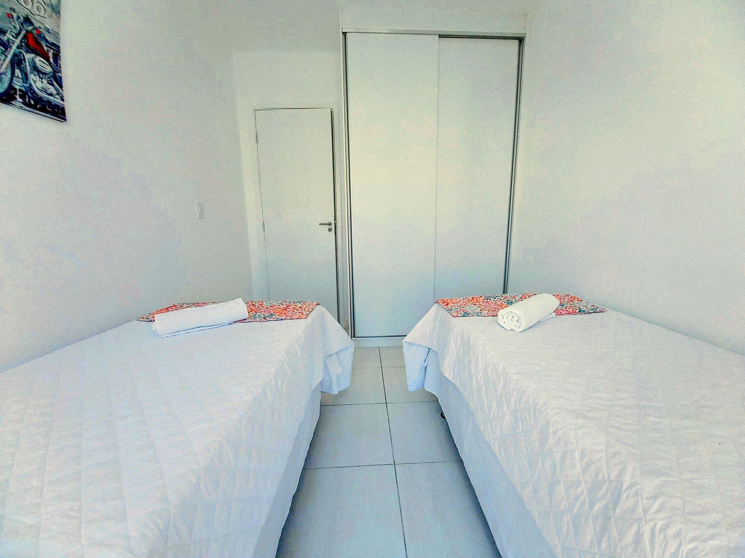 Una habitación con dos camas, cada una con una colcha blanca y una manta estampada con motivos florales. Se observa una puerta al fondo y un cartel en la pared.