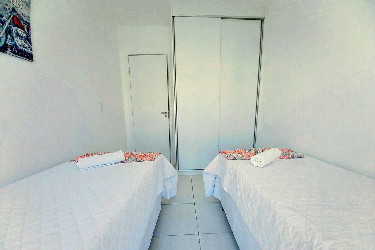 Una habitación con dos camas, cada una con una colcha blanca y una manta estampada con motivos florales. Se observa una puerta al fondo y un cartel en la pared.