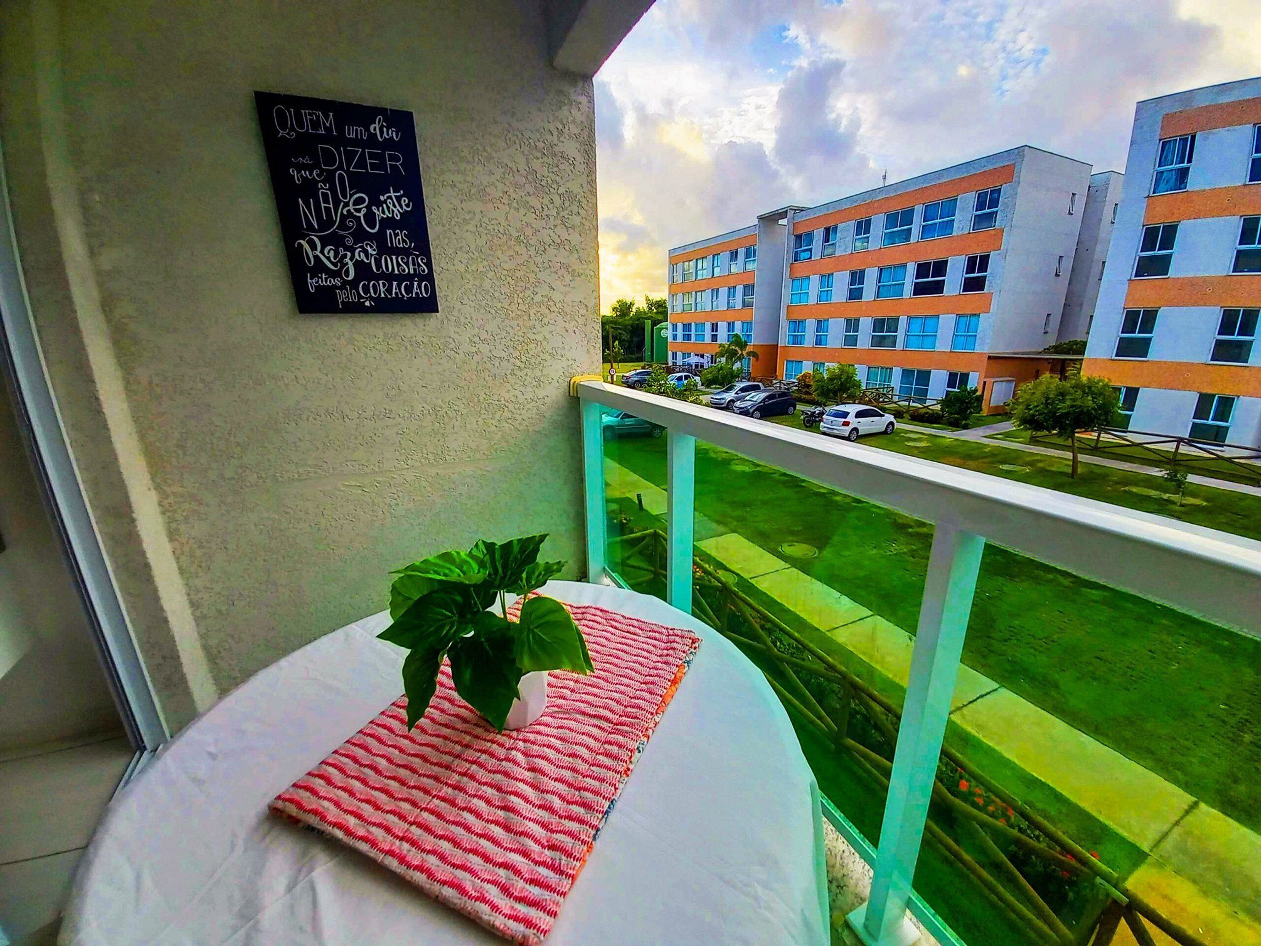 A balcony with a bed, a potted plant, and a view of a modern building with multiple floors and parked cars.