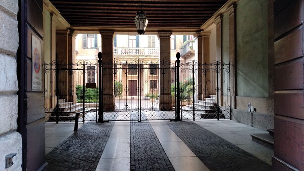 A covered entrance with a wrought iron gate, stone columns, and a tiled floor leading to a courtyard with steps and greenery. - Marconi Rooms - Loggia delle Erbe (Verona)