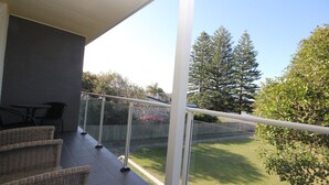 Ein Balkon mit Korbmöbeln, einer Glasreling und Blick auf eine Rasenfläche und Bäume.