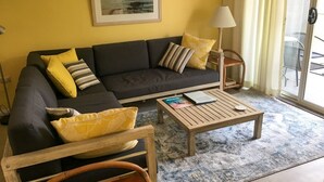 Ein Wohnzimmer mit einem dunklen Sofa, gelben Kissen, einem Holztisch und einem Korbstuhl.