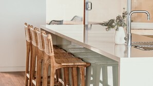 A kitchen with a bar area featuring wooden chairs with woven seats and a stainless steel sink. - Hearns Beachside Villa 9 (Port Fairy)