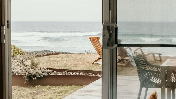 A view from a room with a sliding glass door overlooking a beach with chairs and a table.