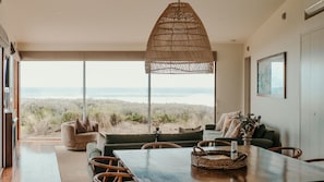 Un vaste espace de vie avec une grande fenêtre donnant sur une plage, une table à manger et un coin salon confortable.