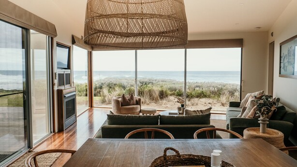 A spacious living area with a large window overlooking a beach, furnished with a sofa, chairs, and a dining table. - Hearns Beachside Villa 9 (Port Fairy)