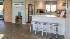 A modern kitchen with a central island, stainless steel appliances, and three bar stools. - NEWLY RENOVATED, LARGE, SERENE, SUNNY  HOME ON PRIVATE ROAD (North Truro)