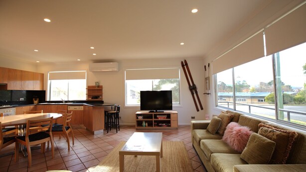A modern kitchen with a dining area, a TV, and a sofa by large windows.