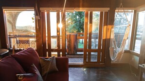 En hyggelig opholdsstue med en rød sofa, et træbord og en hængekøje ved et stort vindue med udsigt til en terrasse og grønne omgivelser.