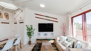 A modern living room with a flat-screen TV, a dining area with a table and chairs, a sofa with decorative pillows, a large window with a view, and a bamboo plant. - Snowdome - 4/3 Poly Cow Lane (Jindabyne)