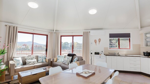A modern living room with a sofa set, a dining table, and a kitchen area with a window and a view of the outdoors. - Snowdome - 4/3 Poly Cow Lane (Jindabyne)