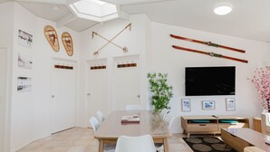 A modern living room with a dining area, featuring a large flat-screen TV, ski equipment, and a bamboo plant. - Snowdome - 4/3 Poly Cow Lane (Jindabyne)