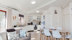 A modern kitchen with white cabinets, a dining area with a glass table, and a cozy seating area with a sofa and cushions. - Snowdome - 4/3 Poly Cow Lane (Jindabyne)