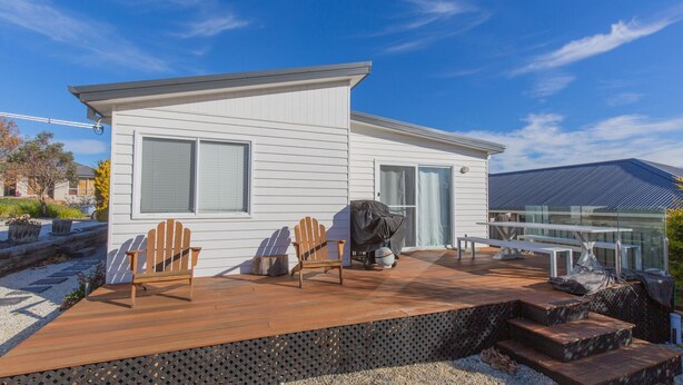 Een modern huis met een houten terras, twee Adirondack stoelen, een barbecue en een glazen patio.