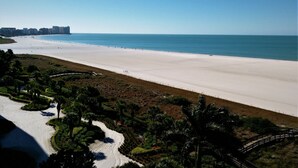 A beach with a clear sky, a sandy shore, and a few people walking. There are buildings in the background and a bridge leading to the beach.