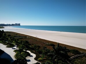 Uma praia com céu claro, areal extenso e algumas pessoas caminhando. Prédios ao fundo e uma ponte que dá acesso à praia.