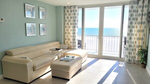 A living room with a beige sofa, a small table, and a chair. There are framed pictures on the wall and a view of the sea through the balcony doors.