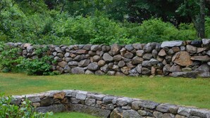 A stone wall in a garden with grass and greenery around it. - Menemsha Home Fully Renovated w/ Amazing Waterview's of Vineyard Sound (Menemsha)