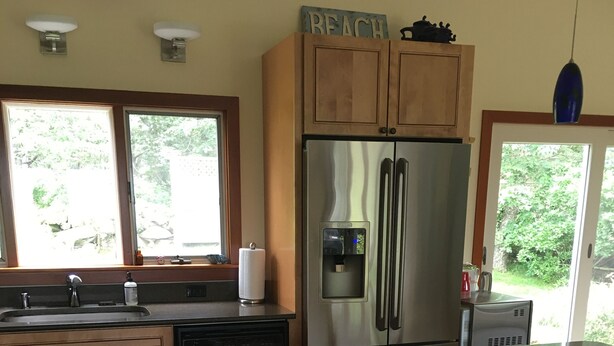 A kitchen with wooden cabinets, a stainless steel refrigerator, a water dispenser, and a window with a view of greenery. - Menemsha Home Fully Renovated w/ Amazing Waterview's of Vineyard Sound (Menemsha)