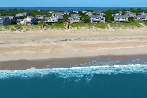 A coastal residential area with houses on a sandy beach, ocean waves, and a clear sky.