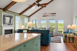 A spacious living room with a high ceiling, a fireplace, and a large window overlooking greenery.