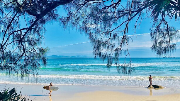 A beach with clear skies, ocean waves, and two people with surfboards.
