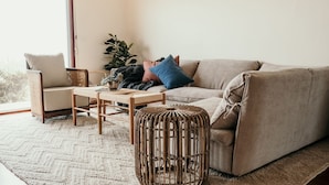 A cozy living room with a beige sofa, a wooden coffee table, a wicker side table, and a large window providing natural light. - Hearns Beachside Villa  7 (Port Fairy)