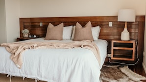A neatly made bed with a white duvet and a beige knitted blanket, a wooden headboard, a bedside lamp, and a small wooden nightstand with a lamp. - Hearns Beachside Villa  7 (Port Fairy)