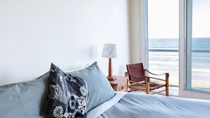 Une chambre à coucher avec un lit, des oreillers, une table de chevet, une lampe et une chaise près d’une grande fenêtre donnant sur l’océan.