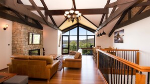 A spacious living room with a stone fireplace, wooden flooring, and a view of the outdoors through large windows. - Heidelburg - 3/6 Banjo Patterson Crescent (Jindabyne)