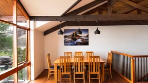 A dining area with a wooden table and chairs, a large window, and a black pendant light. - Heidelburg - 3/6 Banjo Patterson Crescent (Jindabyne)