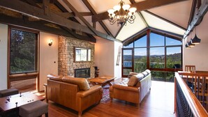 A spacious living room with a stone fireplace, leather sofas, and a large window offering a scenic view. - Heidelburg - 3/6 Banjo Patterson Crescent (Jindabyne)