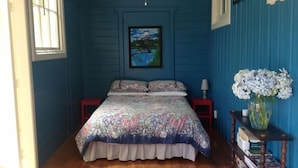 A bedroom with a floral bedspread, a nightstand with a lamp, a framed picture on the wall, and a bookshelf with books and a vase of flowers.