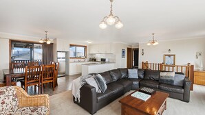Ein modernes Wohnzimmer mit einem dunkelbraunen Ledersofa, einem Holzesstisch und Stühlen. Es gibt eine Küchenzeile mit weißen Schränken und eine Kochinsel. Das Zimmer verfügt über ein großes Fenster und eine Chandelüster.