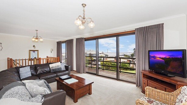 Ein großzügiges Wohnzimmer mit einer breiten Couch, einem Couchtisch und einem Fernseher auf einem Holzständer. Das Zimmer verfügt über einen Balkon mit Blick auf die Umgebung und eine große Schiebeglastür.