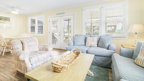 Un salón luminoso con muebles a rayas, una cesta de mimbre sobre una mesa de madera y vistas a través de la puerta.