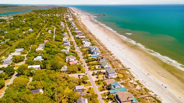 An aerial view of a coastal residential area with houses along the beach, a pier extending into the ocean, and clear skies. - Blue Sky | Two Units | Pool w/Heating Option | Across from Beach Access (Folly Beach)