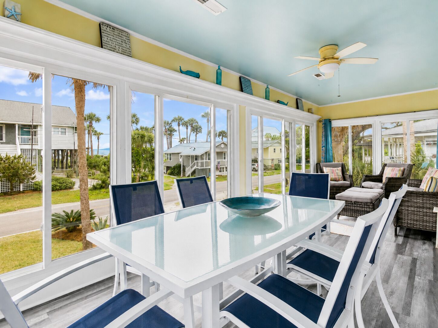 A sunlit room with a glass table, white chairs, and a view of the neighborhood.