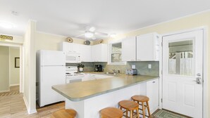 A kitchen with white cabinets, a central island with stools, a ceiling fan, and a caution sign on the wall. - Blue Sky | Two Units | Pool w/Heating Option | Across from Beach Access (Folly Beach)