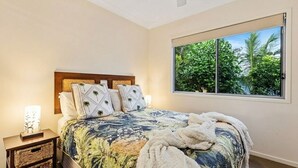 A bedroom with a large bed, a nightstand, a lamp, and a window with a view of greenery. - 15 Esprit Drive - Rainbow Shores. Wifi. Close to beach (Rainbow Beach)